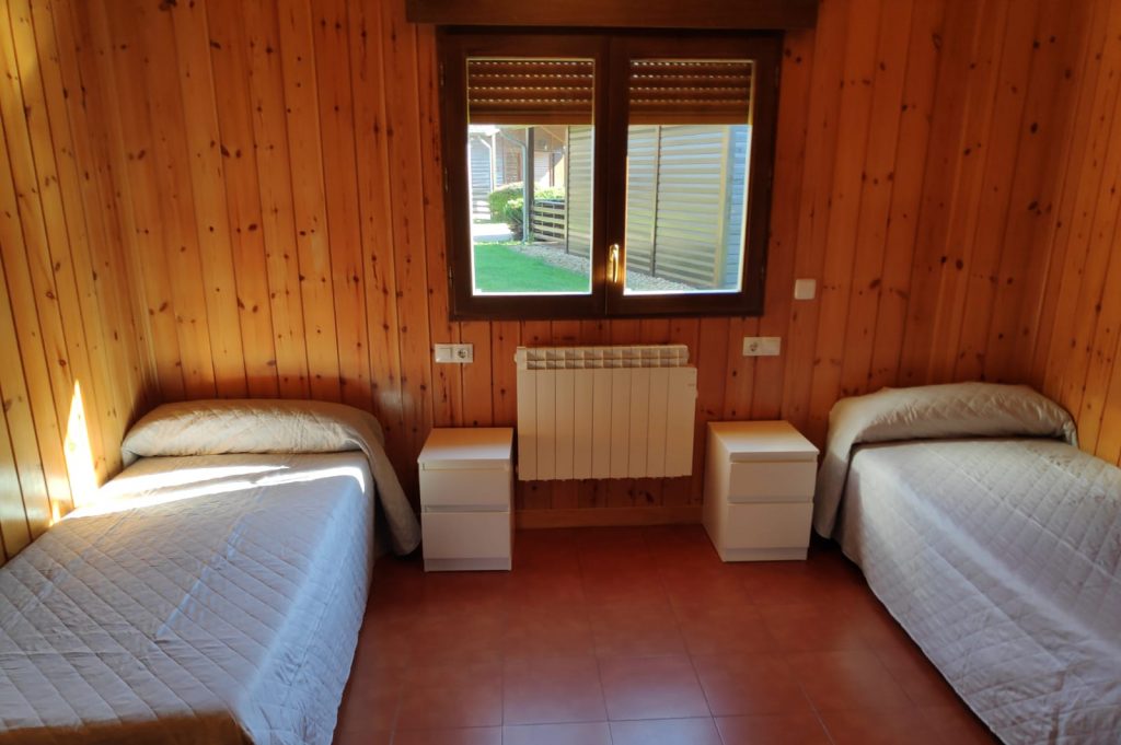 Dos camas y dos mesillas, con un radiador y una ventana entre ambas